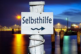Auf einem Schild angebracht an einen Pfeiler steht das Wort "Selbsthilfe" geschrieben. Dazu ein Pfeil nach rechts. Im Hintergrund ist ein Hafenbecken mit Schiffen vor einem frühabendlichen Himmel zu sehen. 