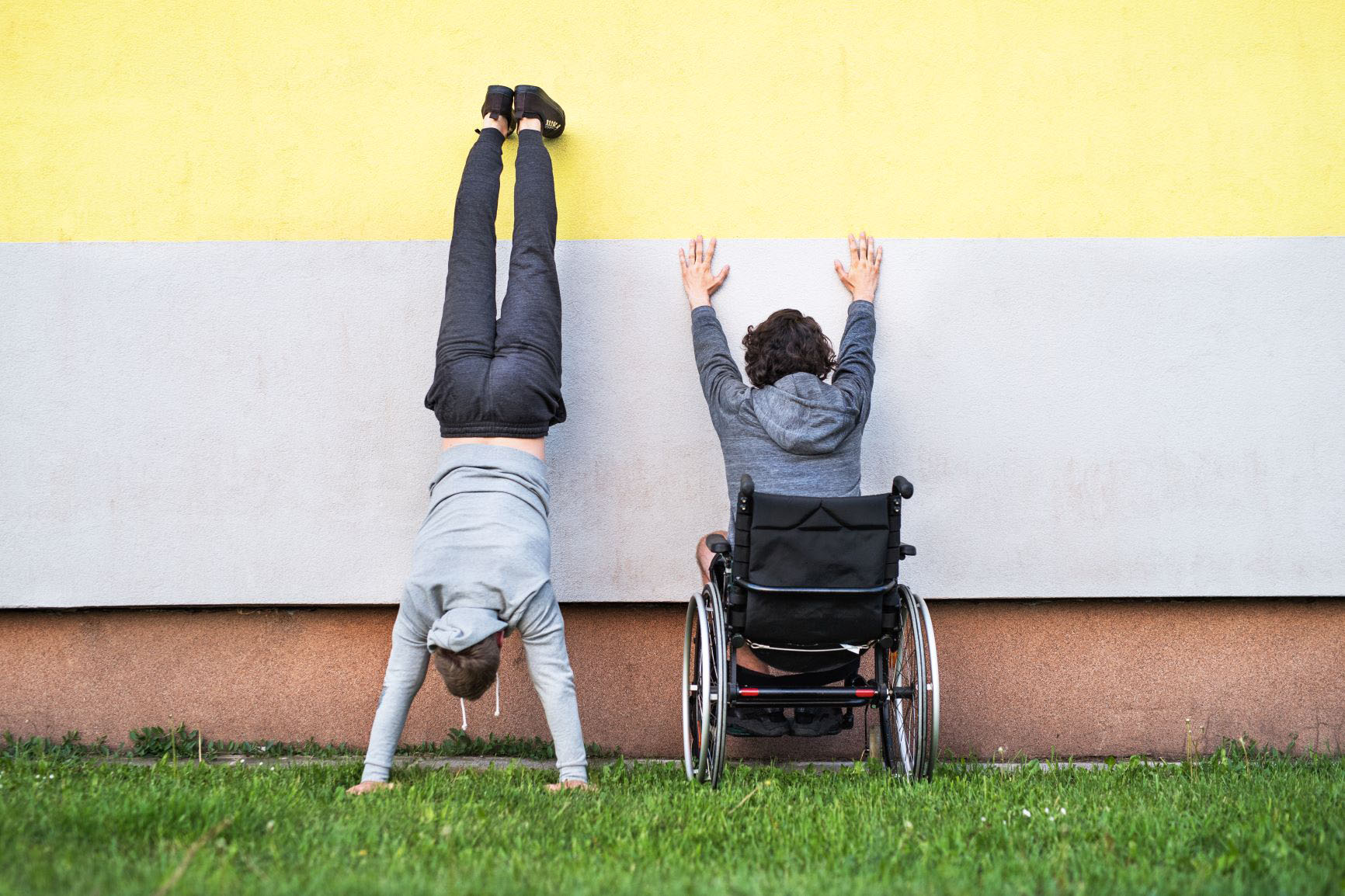 Ein Mensch mit und ein Mensch ohne Behinderung vor einer bunt gestrichenen Hauswand 